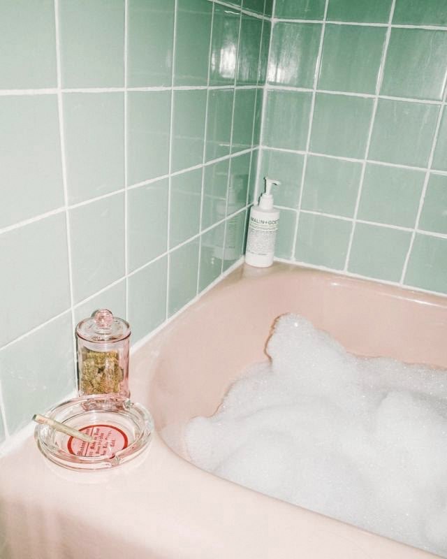 A pale pink bathtub full of bubbles against mint green tiles, with a glass jar and ashtray on the edge